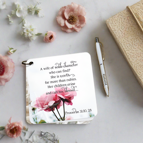 Scripture card with the Bible verse of Proverbs 31:10,28 printed over a picture of blooming pink roses. The  bible verse cards are laying on a white background surrounded by flowers a pen and a book.