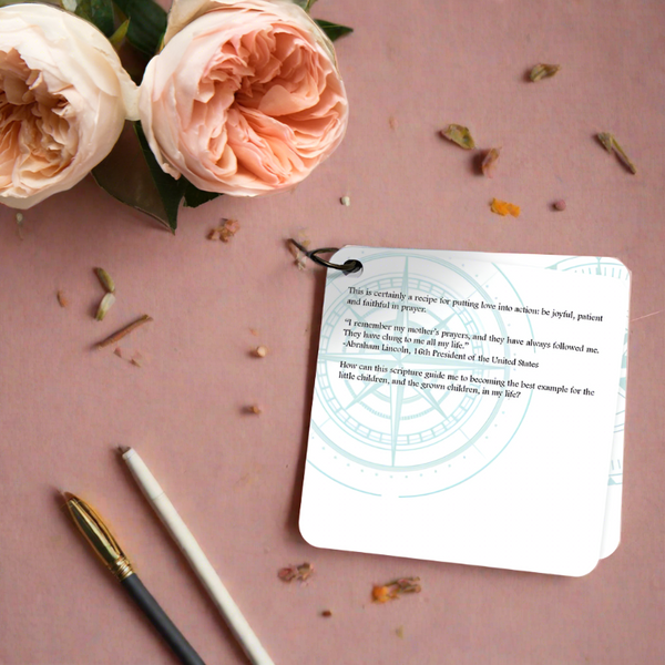 A scripture devotional card printed on a white background with a compose rose etch in teal. the devotion is laying on a neutral background with two pale pin roses and a pen & pencil laying beside it. 