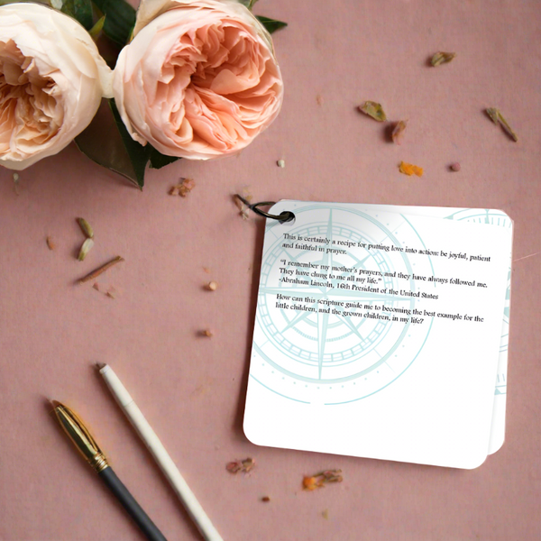 A scripture devotional card printed on a white background with a compose rose etch in teal. the devotion is laying on a neutral background with two pale pin roses and a pen & pencil laying beside it.