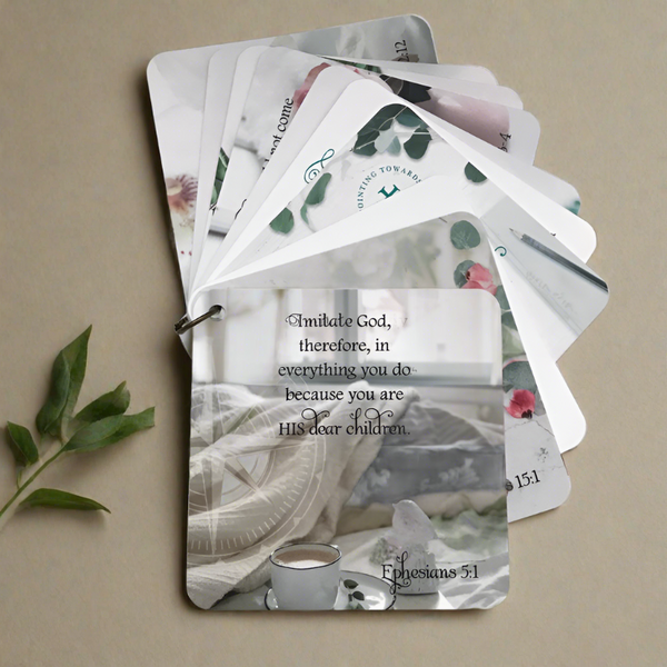 Scripture card of Ephesians 5:1  Bible verse printed over a photo of an unmade bed with a cup of coffee sitting on a saucer that is sitting on a bed in front of an open window. The card is fanned out on top of other cards in the set, on a neutral background with green leaves laying beside them. Bible verse gift idea. I
