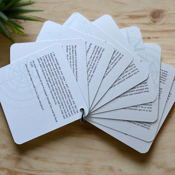 The backside of the scripture cards fanned out. Each card has a devotion printed on it . They are laying on a plywood background with greenery around the top.