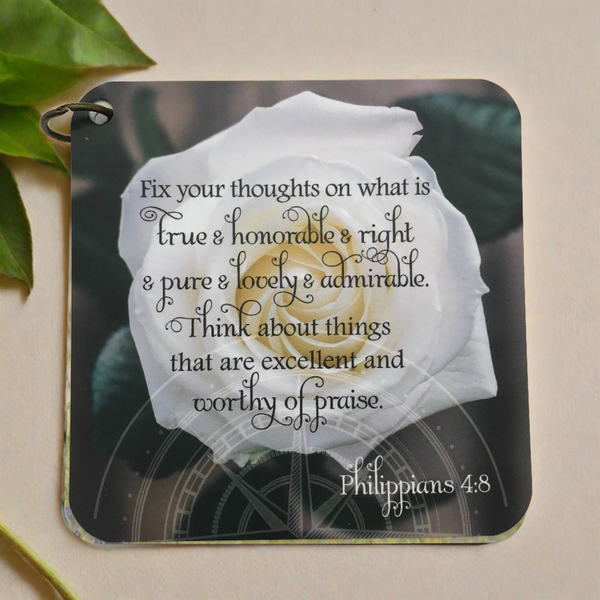 Bible verses card with the scripture Philippians 4:8 printed over a photo of a blooming white rose.  Laying on a neutral background with green leaves at the top left corner. Encouraging bible verses. Mother's Day gift idea.