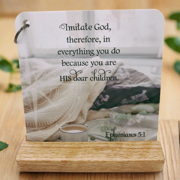 Scripture card of Ephesians 5:1 is printed over a photo of an unmade bed with a cup of coffee sitting on a saucer that is sitting on a bed in front of an open window. Card is displayed on an oak easel with blurred out greenery  in the background.