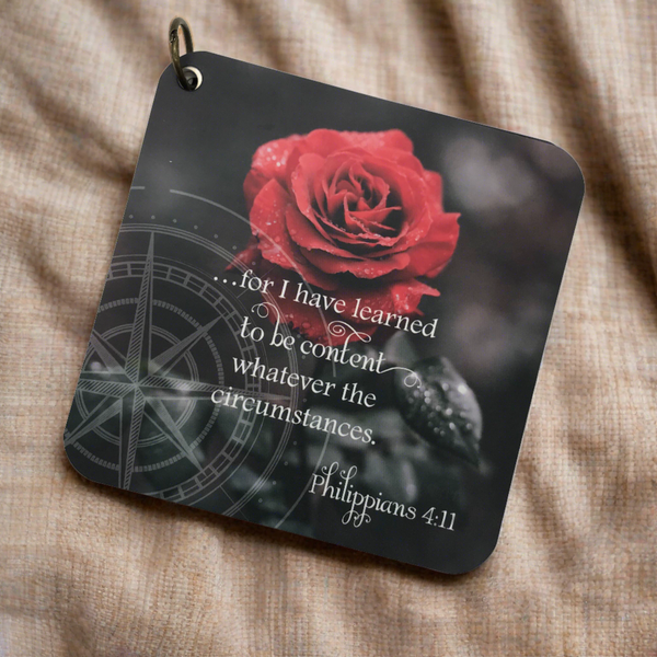 A scripture card with the bible verse Philippians 4:11 printed over a black and white background with a blooming read rose in the background. The scripture devotional card is laying on a piece of burlap fabric.