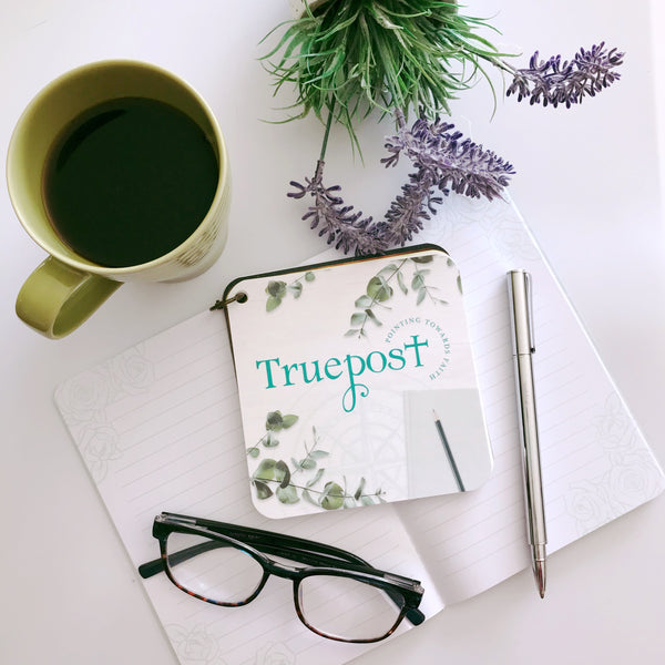 Cover card for a scripture devotional card set. The cover says Truepost and has a compass rose etched in the background. The cards are sitting with a coffee cup, a journal, reading glasses, pen and spring flowers.