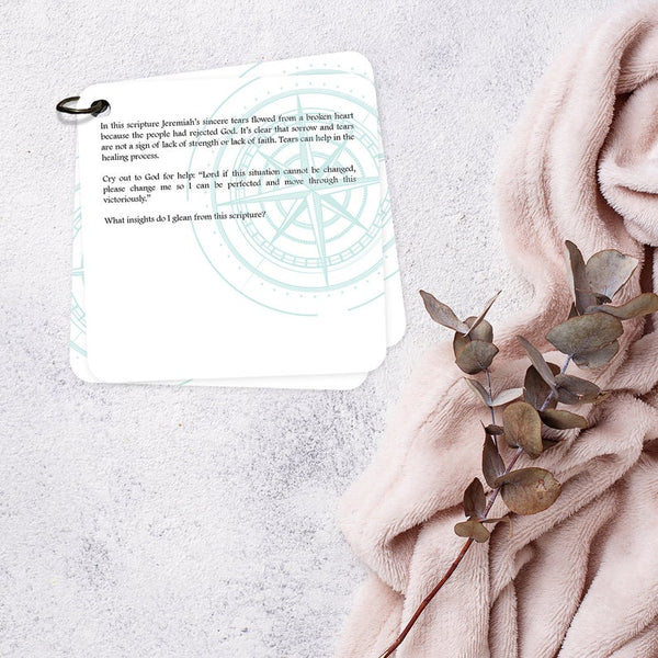 The backside of one of the scripture devotional cards with a devotion printed on it. The cards are laying on a cream blanket with another cream colored blanket and greenery laying beside it.