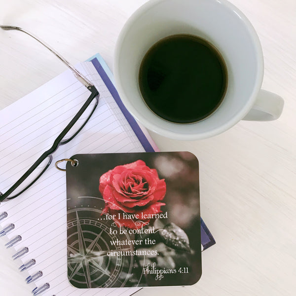 Motherhood scripture card / bible verse with Philippians chapter 4 verse 11 printed over  a black and white background with a bright red rose. The inspirational bible verse card is laying on top of a small lined notebook next to eyeglasses and a cup of coffee to the side.