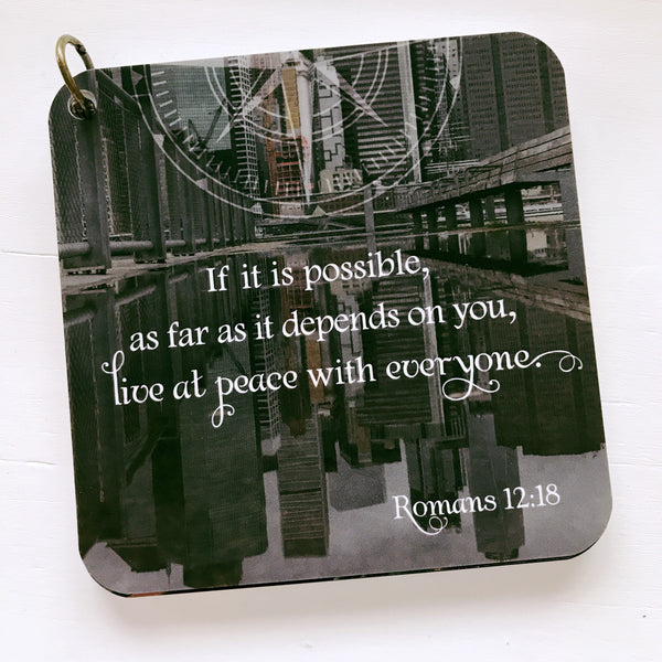Scripture card of Romans 12:18 printed over a black and white photo of a city scape reflecting off a pool.