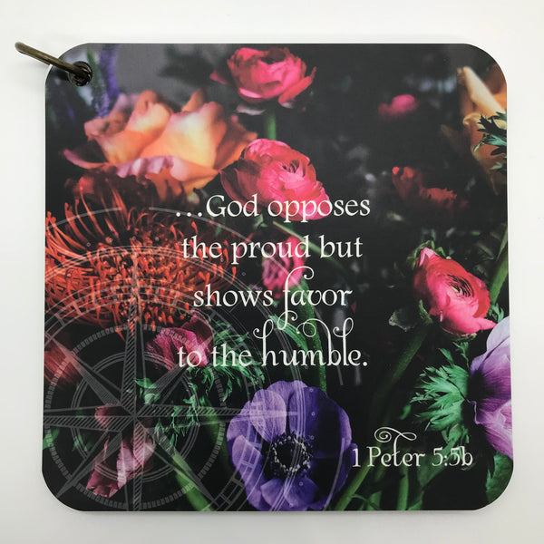 A scripture devotional card of 1Peter 5:5b printed over a photo of multicolored spring flowers. A compass rose is etched in the photograph.