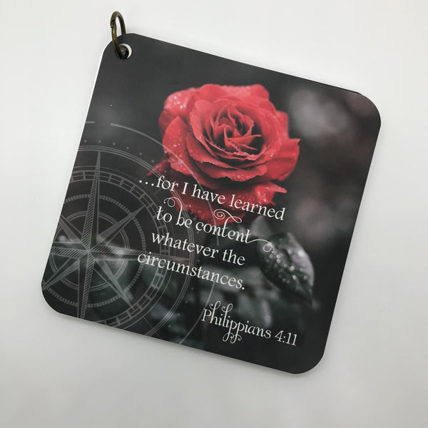 A scripture devotional card of Philippians 4:11 printed over a photograph of a black and white background of a red rose. There is also a compass rose etched in the photo.