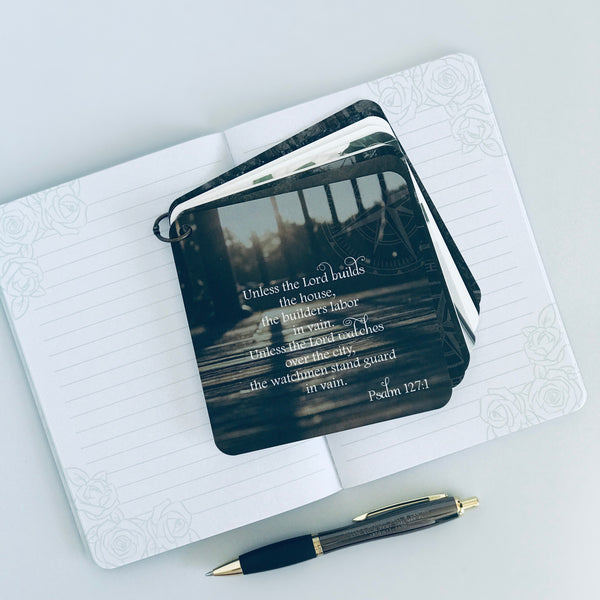 Scripture card of Psalm 127:1 is printed over a photo of what looks like the rod iron fence railing around a wooden deck, with the sun peeking through the rails. The card is laying on an open page of a lined journal.