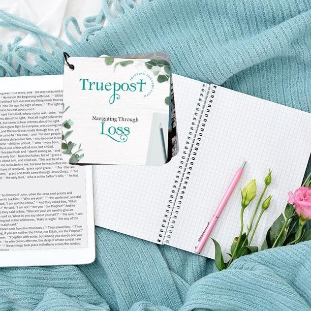 Scripture devotional card set called navigating through loss. It's pictured laying on top of a turquoise blanket and notepad, along with an iPad and pink roses.