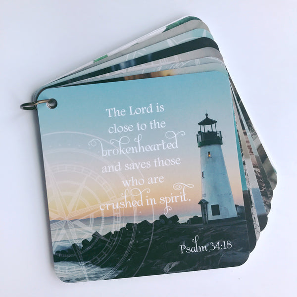 Scripture card on loss and broken heartedness from Psalm 34:18 is printed over a photo of a lighthouse sitting on rocks at the edge of the water with a setting sun in the background. It's laying on a white background.