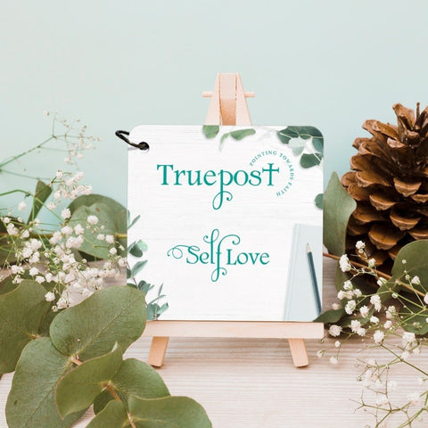 Truepost pointing towards faith self love cover card with eucalyptus. Sitting on an easel next to pine cones and greenery.
